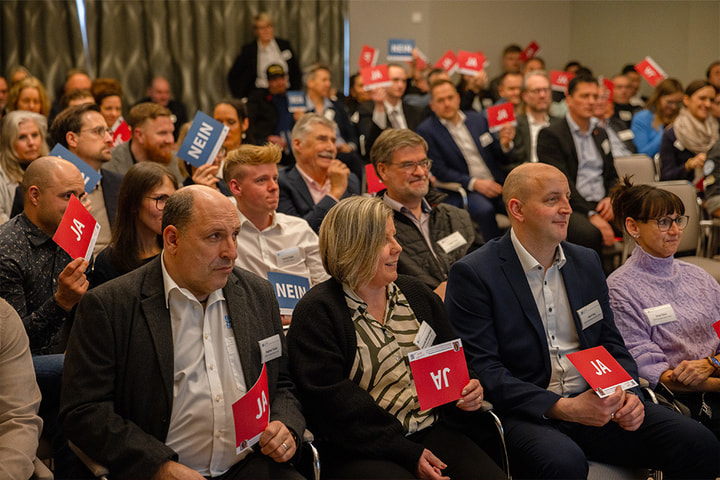 Publikum bei einer Abstimmung in einem Saal: Mehrere Personen halten rote „JA“- und blaue „NEIN“-Karten hoch.