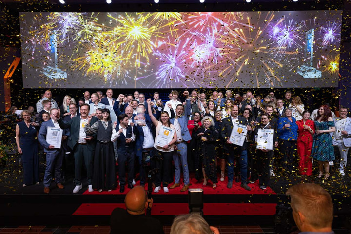 Eine große Gruppe jubelnder Preisträgerinnen und Preisträger steht auf einer Bühne, während goldene Konfetti fällt und im Hintergrund bunte Feuerwerke auf einer Leinwand zu sehen sind.